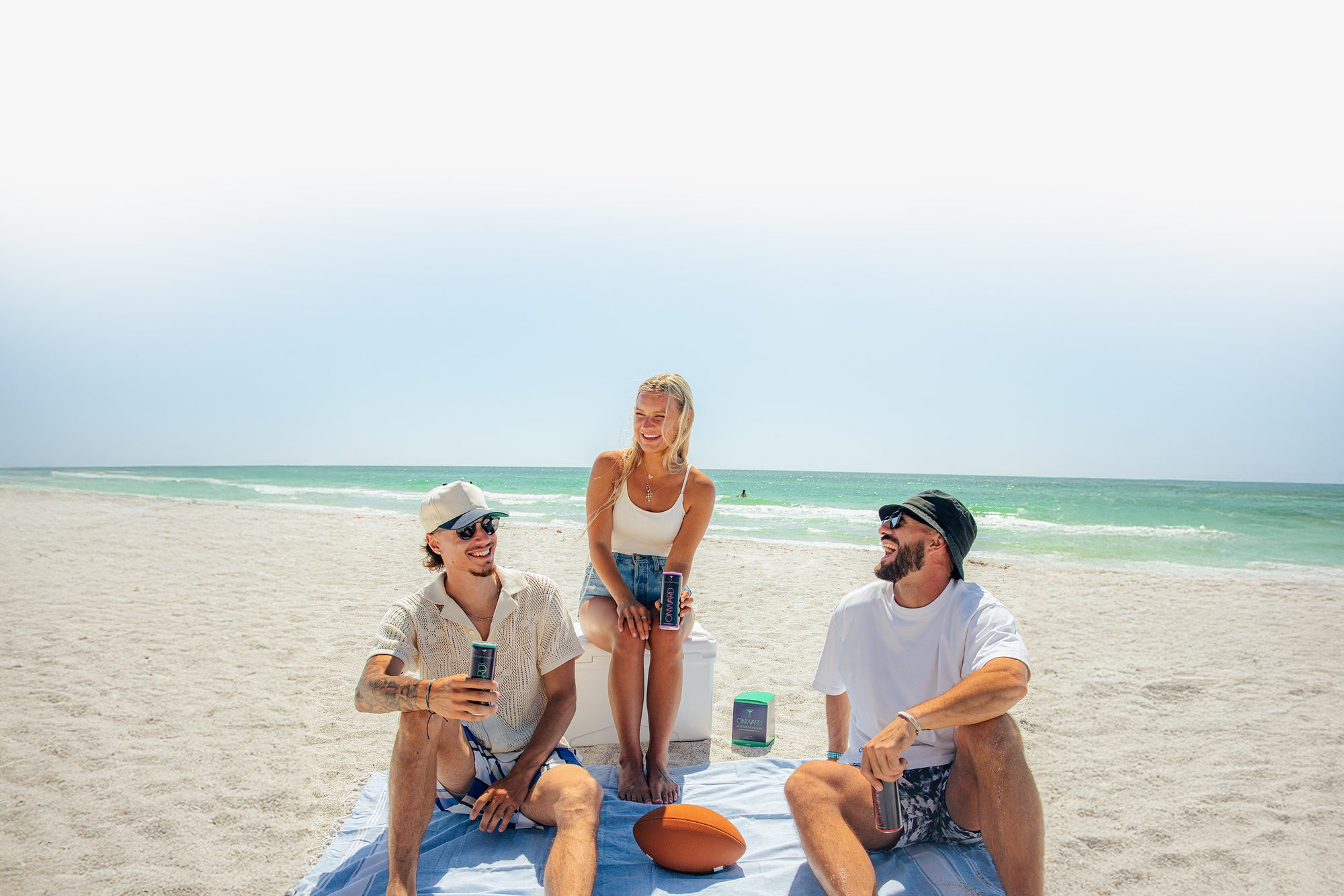 Three people sitting on a towel at the beach with a clear sky and ocean in the background.