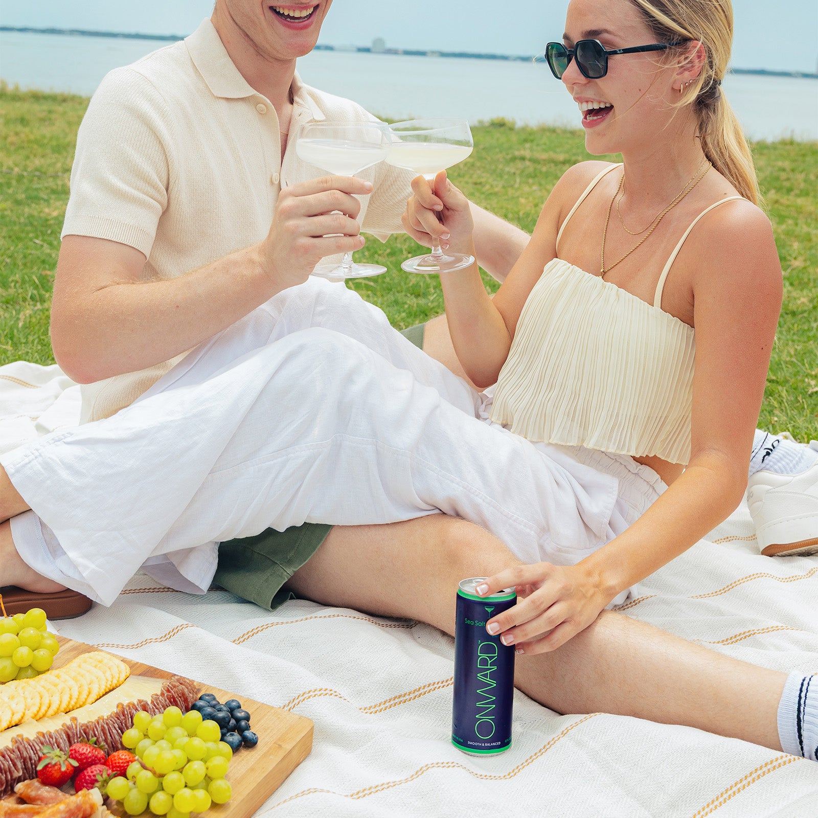 Couple having a picnic by the water, holding drinks and a can of 'Onward' drink.