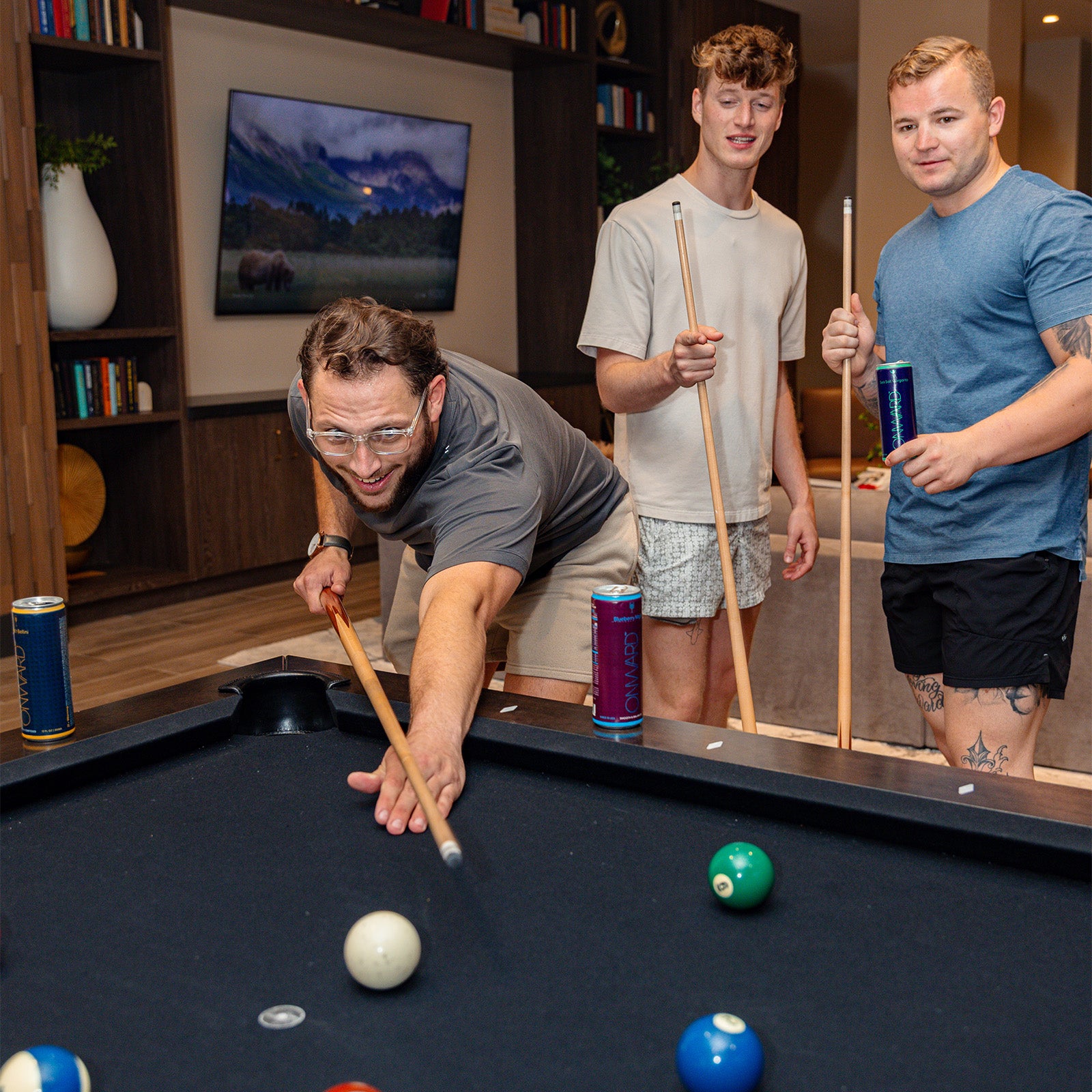 Three men playing pool in a living room with a TV and bookshelf in the background.