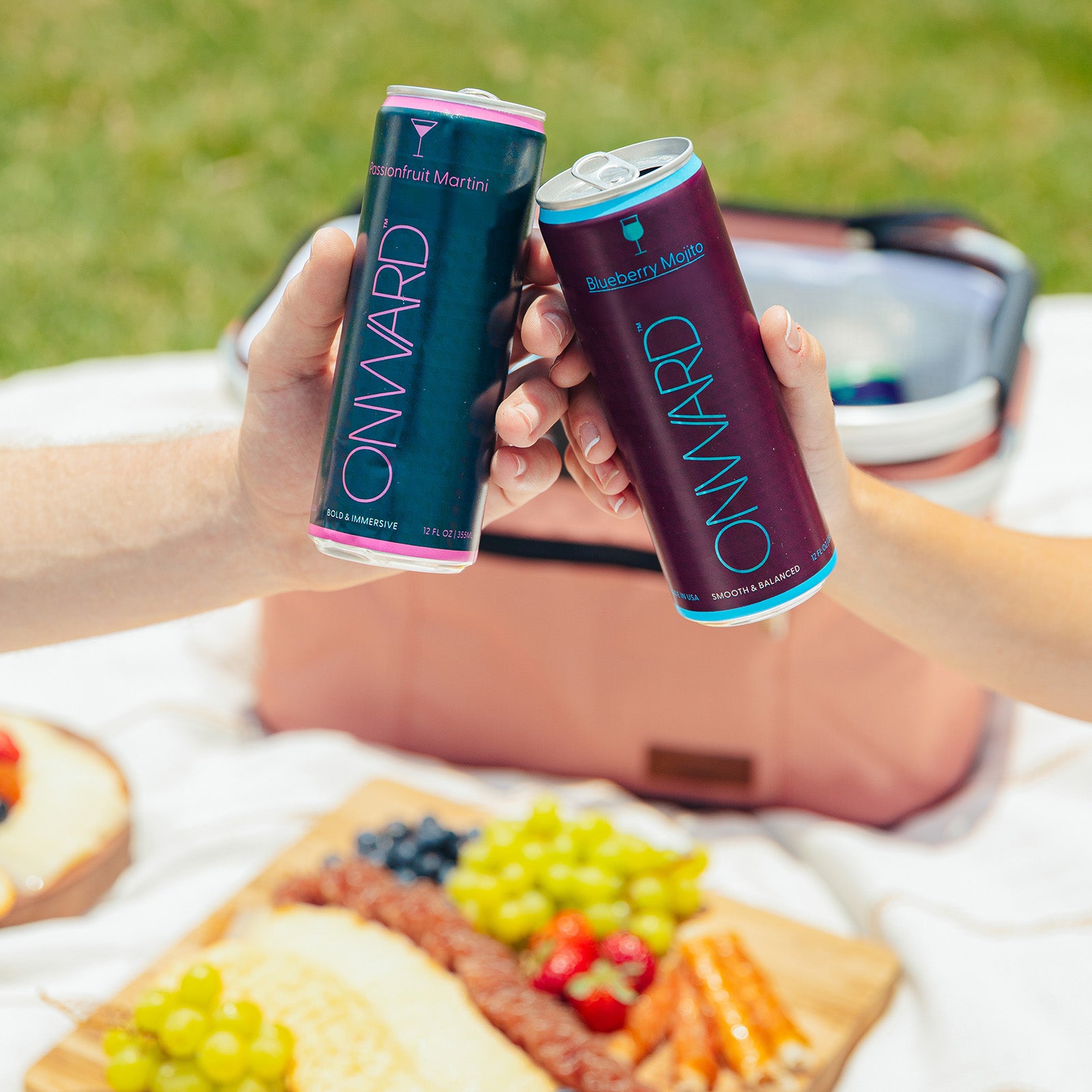 Two cans of 'Onward' drinks held by hands over a picnic setup with fruit and bread.